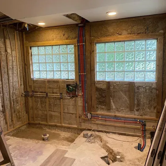 Bathroom under construction, with exposed framing, plumbing, glass block windows, and recessed lighting.